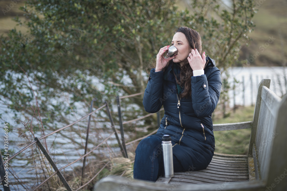 A woman sitting on a wooden bench near a tree overlooking a lake drinks tea from a flask while looking away before sunset in the Pentland hills regional park in Edinburgh, Scotland, United Kingdom A woman sitting on a wooden bench near a tree overlooking a lake drinks tea from a flask while looking away before sunset in the Pentland hills regional park in Edinburgh, Scotland, United Kingdom