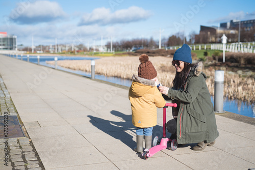 Billede på lærred An Asian mother wearing a wooly hat helps her daughter get ready to ride her sco
