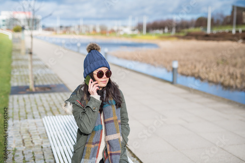 Asian woman wearing a woolly hat and sunglasses talks on her mobile phone while looking away and sitting on a white bench near a Canal in the city of Edinburgh, Scotland, United Kingdom