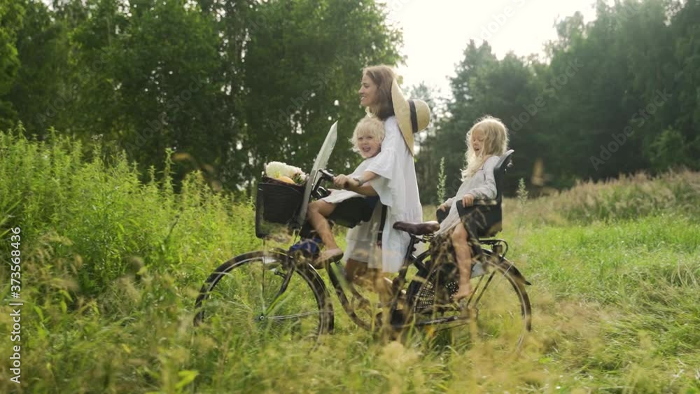 Young beautiful happy mother carries two children on a bicycle on the edge of the forest Young beautiful happy mother carries two children on a bicycle on the edge of the forest