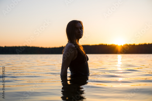 Beautiful Russian girl in a long black swimsuit swims outside the city on the lake in the rays of sunset or dawn. Splashing water on the background of the sun. Happy girl. © Alexey Kartsev