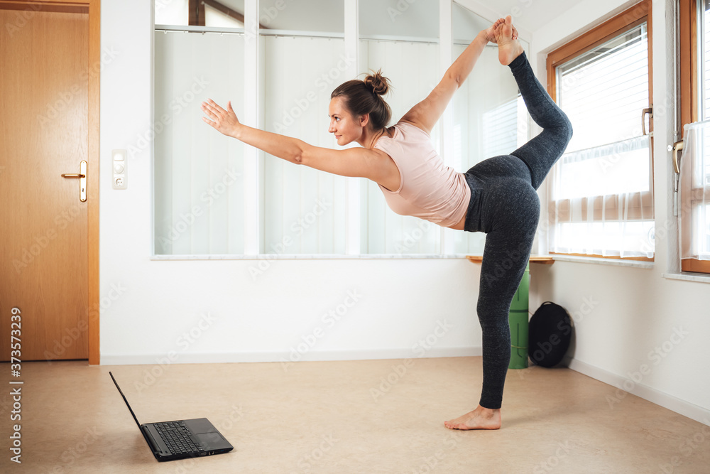Full length of sporty young woman standing on one leg practicing yoga with laptop at home