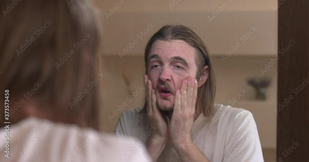 Young man looks at himself in the bathroom mirror, washes face with water Young man looks at himself in the bathroom mirror, washes face with water