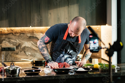 Tattooed chef preparing steak records online broadcasts. Live streaming concept. Interview video recording and stream. Concept of cooking of meat by tattoed butcher vlogging live feeds on social media