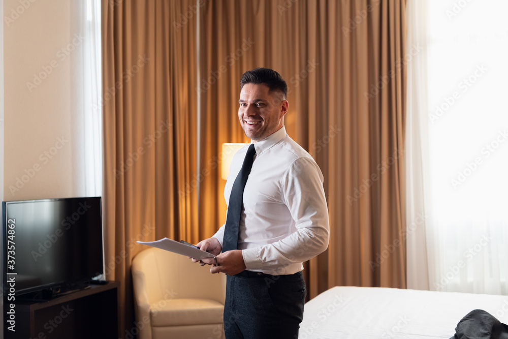 Fototapeta premium A young happy traveler businessman wearing a white shirt and a necktie standing in a hotel room. A smiling businessman with a smartphone and some blueprints in his hands getting ready for a new big