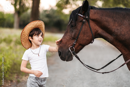 Fototapeta Cute little boy in a cowboy hat stroking a beautiful horse outdoors