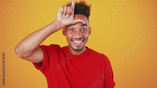 Funny african american guy showing loser gesture and giggling, orange background