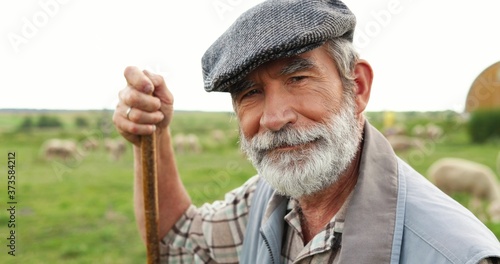 Fotografie Portrait of Caucasian senior handsome man shepherd in hat standing outdoor, leaning on stick, looking at side and turning to camera