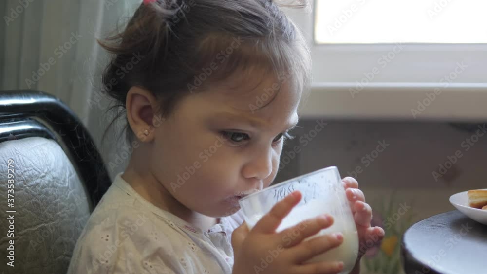 Adorable little girl is eating cheesecakes with her hands at the table in kitchen. Glass of yogurt on the table Adorable little girl is eating cheesecakes with her hands at the table in kitchen. Glass of yogurt on the table