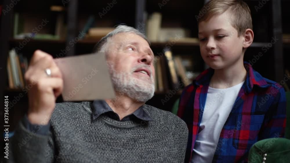 Handsome bearded gray-haired grandfather with his cute grandson having fun looking at an old photo album, enjoying memories watching family photo album at home in cozy room background on bookshelves. Handsome bearded gray-haired grandfather with his cute grandson having fun looking at an old photo album, enjoying memories watching family photo album at home in cozy room background on bookshelves.