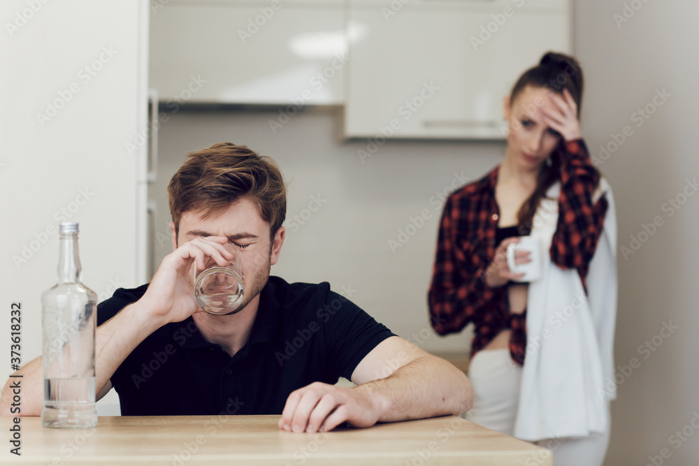 Drinking problem drunk man in a young family. A man drinks while sitting at a table in the kitchen, an upset woman stands next to a man and looks at him Drinking problem drunk man in a young family. A man drinks while sitting at a table in the kitchen, an upset woman stands next to a man and looks at him