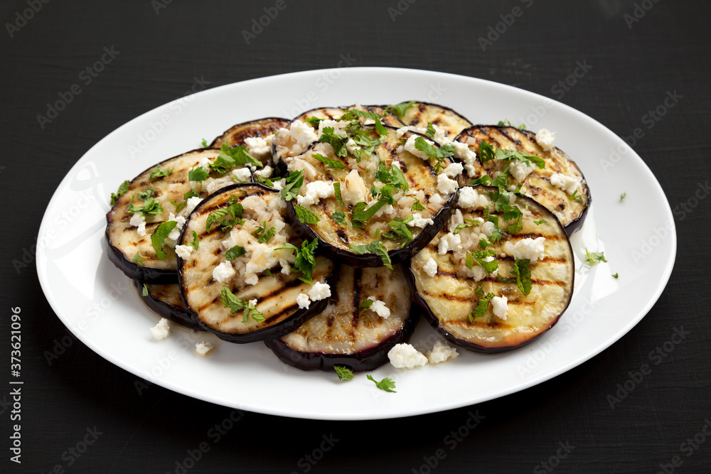Homemade Grilled Eggplant with Feta and Herbs on a white plate on a black surface, side view. Homemade Grilled Eggplant with Feta and Herbs on a white plate on a black surface, side view.