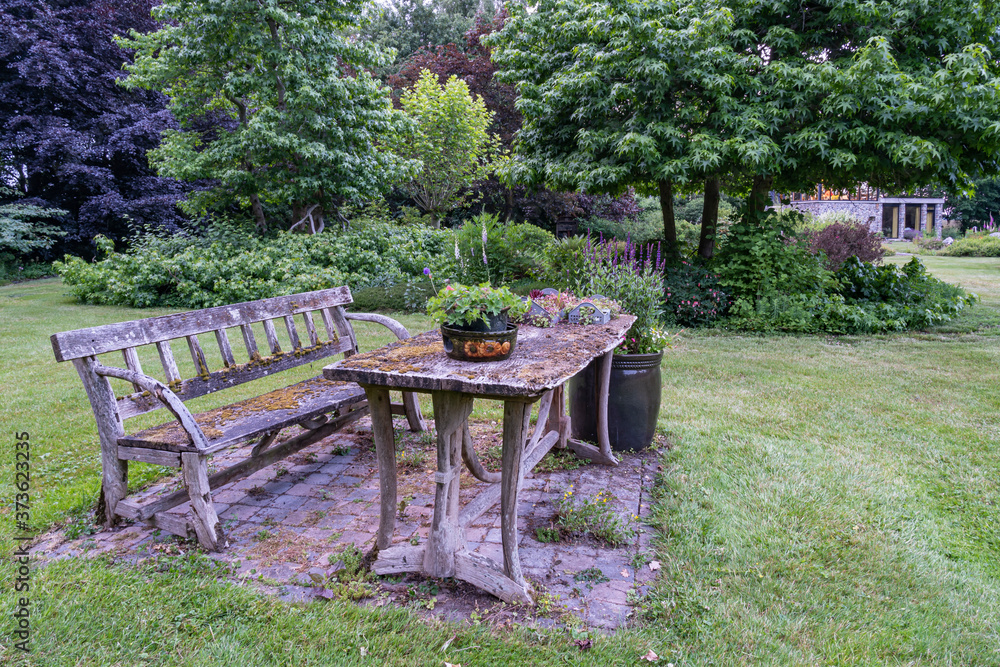 This old wooden bench and table fit perfectly into this color palette of trees and plants in this beautifully landscaped garden with a great diversity of trees and flowering shrubs This old wooden bench and table fit perfectly into this color palette of trees and plants in this beautifully landscaped garden with a great diversity of trees and flowering shrubs