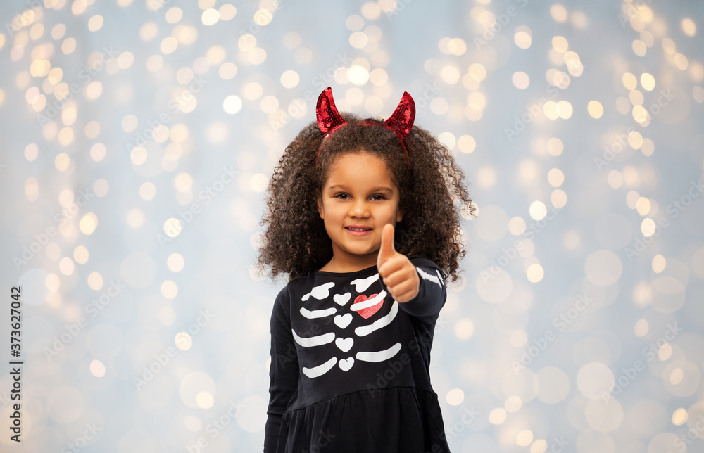 halloween, holiday and childhood concept - smiling african american girl in black costume dress and red devil's horns over festive lights background halloween, holiday and childhood concept - smiling african american girl in black costume dress and red devil's horns over festive lights background