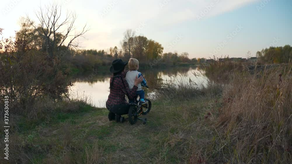 Mother and Son Hiking on Bike Near River. Child Discovers World Mother Tells about Nature Environment. Sunset over Countryside Rural Scene. Slow motion Mother and Son Hiking on Bike Near River. Child Discovers World Mother Tells about Nature Environment. Sunset over Countryside Rural Scene. Slow motion