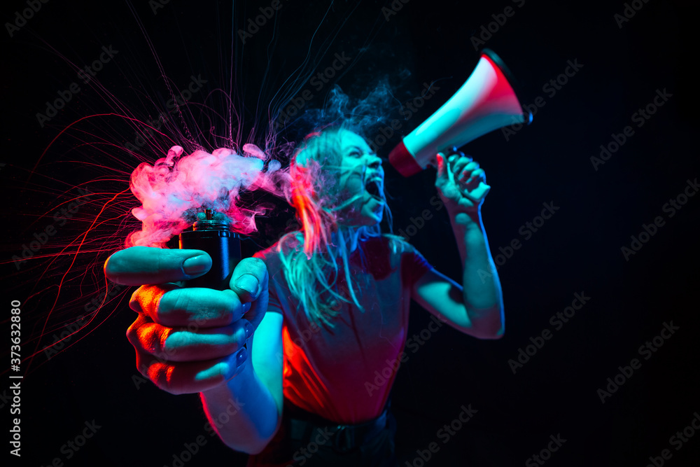Shouting with megaphone. Young woman with smoke and neon light on black background. Highly tensioned, wide angle, fish eye view. Concept of human emotions, facial expression, sales, ad, sport.