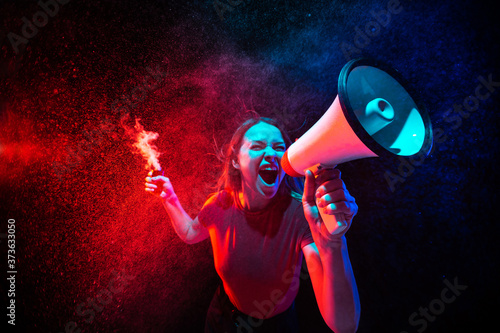 Shouting with megaphone. Young woman with smoke and neon light on black background. Highly tensioned, wide angle, fish eye view. Concept of human emotions, facial expression, sales, ad, sport.