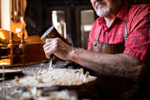 An experienced senior carpenter holding knife and hammer carving wooden board in his woodworking workshop.