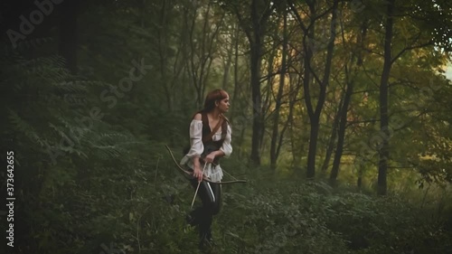 Medieval woman walks in the forest. A girl holds a wooden bow in her hands. Fantasy image of Lady hunter. Vintage warrior archeress. Creative clothes leather pants, white shirt jacket, bag with arrows