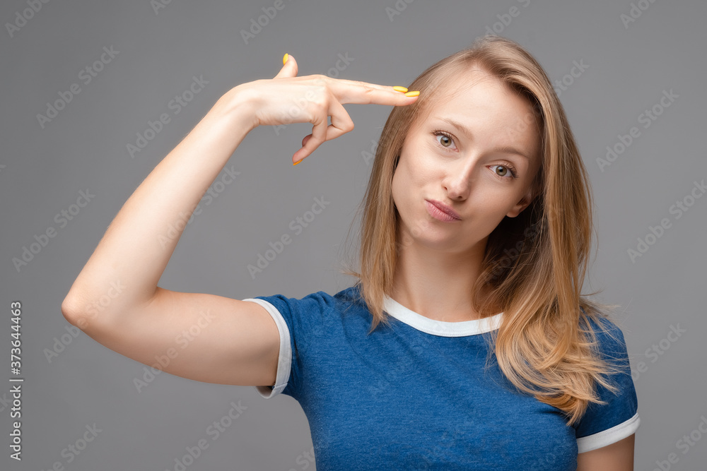Funny young blonde female shoots in temple, tilts head, dressed in casual blue t shirt, demonstrates suicide gesture Funny young blonde female shoots in temple, tilts head, dressed in casual blue t shirt, demonstrates suicide gesture
