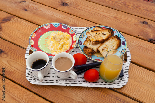 Healthy breakfast tray with pinneapple juice, cofee cups, tomatos, scrambled eggs and bread toasts with cuttlery on a wooden background outdoors table. Nutritious breakfast concept with negative space photo