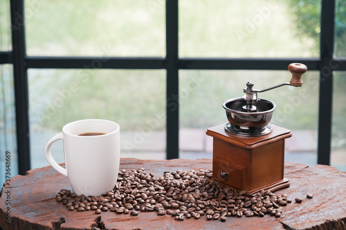 fresh coffee in white cup with grinder and roasted seed on wood table with blurred black windows frame and green garden home's yard in background