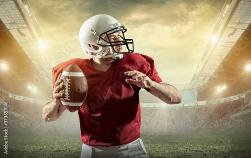 American football sportsman player with ball in action on stadium under lights of background. Sport, proud footballer in white helmet and red t-shirt ready to play.