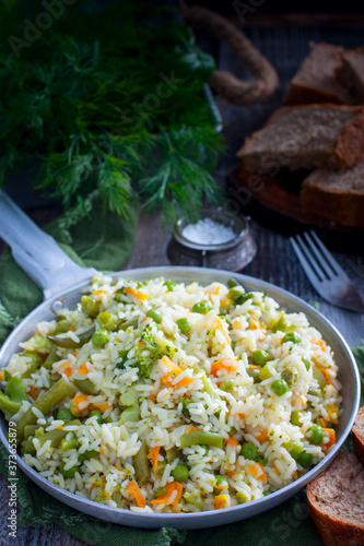 Rice with vegetables - carrots, onions, broccoli, pepper, green peas in a frying pan on a wooden table, selective focus