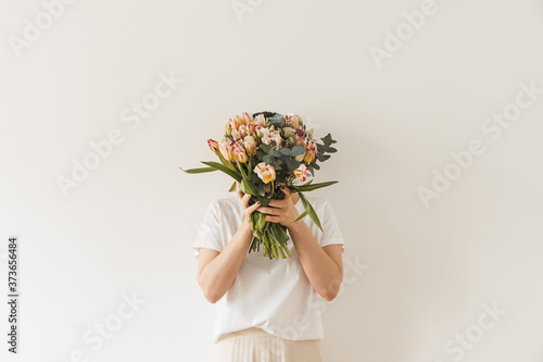 Fototapeta Naklejka Na Ścianę i Meble -  Young pretty woman in white blouse holding tulip flowers bouquet in hands against white wall. Holiday celebration festive floral concept