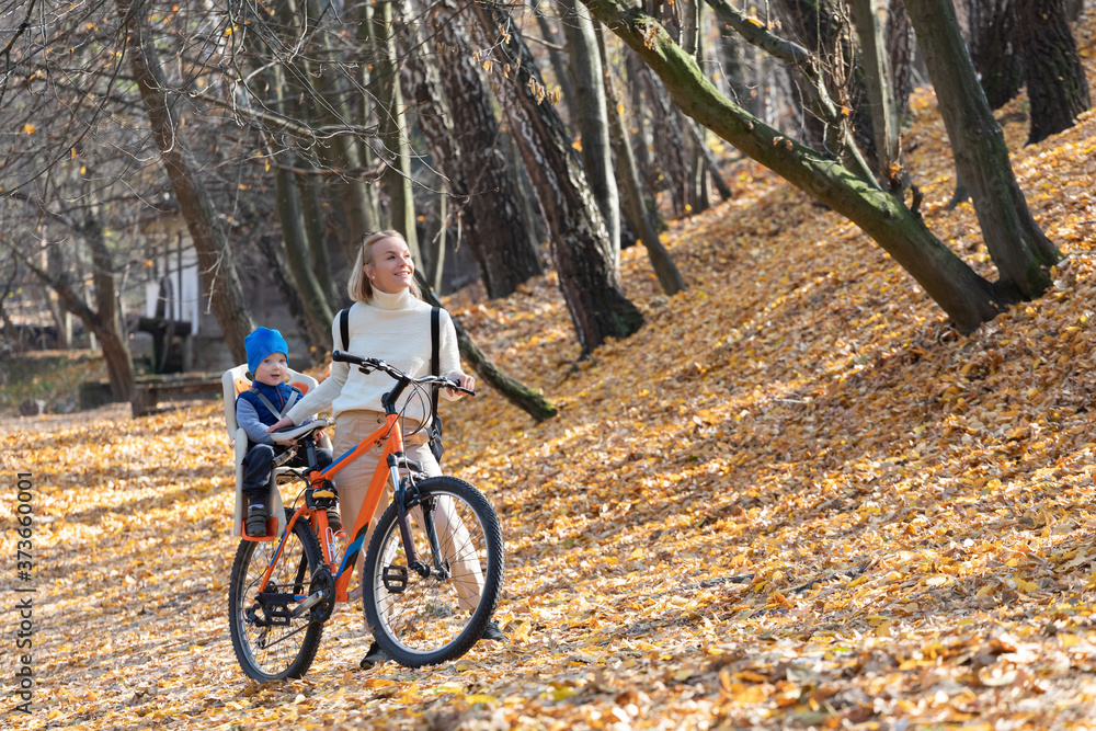 Fototapeta premium Happy mother leads a bicycle with a child strapped in the back in the autumn park.