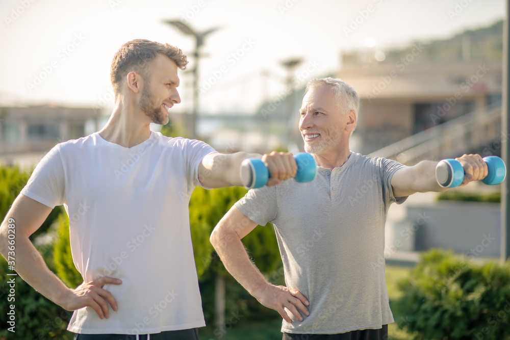 Bearded male and mature man holding blue dumbbells, exercising, smiling Bearded male and mature man holding blue dumbbells, exercising, smiling