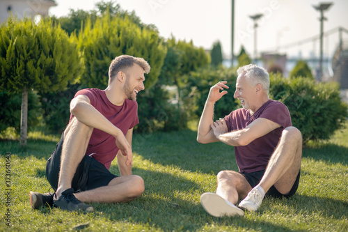 Bearded male and grey-haired man sitting on grass, relaxing after workout