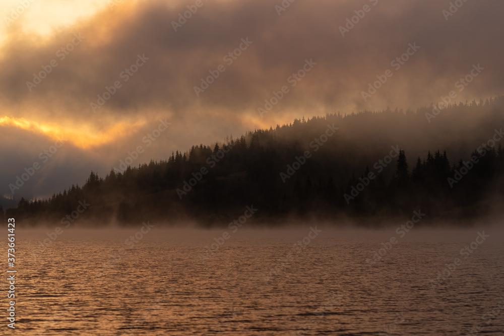 Dramatic view of a natural landscape in a mountainous area with a quiet lake and a coniferous forest shrouded in fog with the sun rising behind the clouds in the early hours of the morning. Dramatic view of a natural landscape in a mountainous area with a quiet lake and a coniferous forest shrouded in fog with the sun rising behind the clouds in the early hours of the morning.