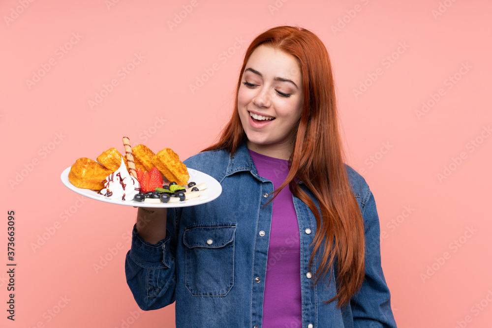 Redhead teenager girl holding waffles over isolated pink background with happy expression