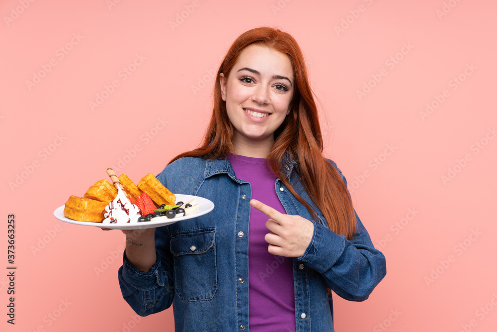 Redhead teenager girl holding waffles over isolated pink background and pointing it