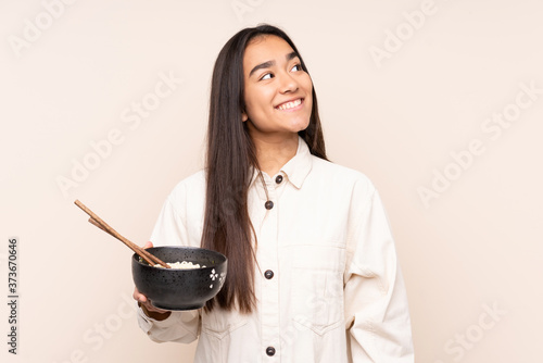 Young Indian woman isolated on beige background looking up while smiling while holding a bowl of noodles with chopsticks