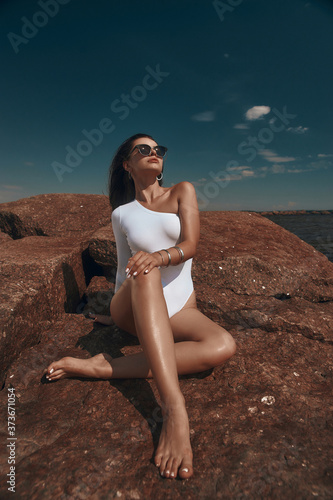 High fashion oytdoor portrait. Sexy tanned caucasian woman with long brunette straight hair in white body and sunglasses sitting at huge stone or rock at seacoast on a sunny day