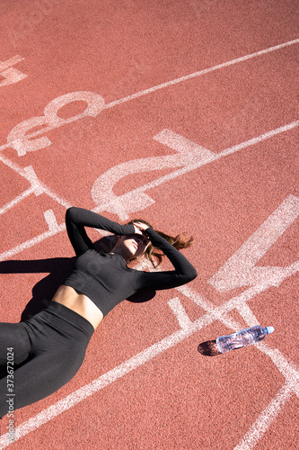 Top view of tired fit woman in sportswear resting after workout or running on a treadmill rubber stadium, holding a bottle of water, taking a break during training, outdoors. 