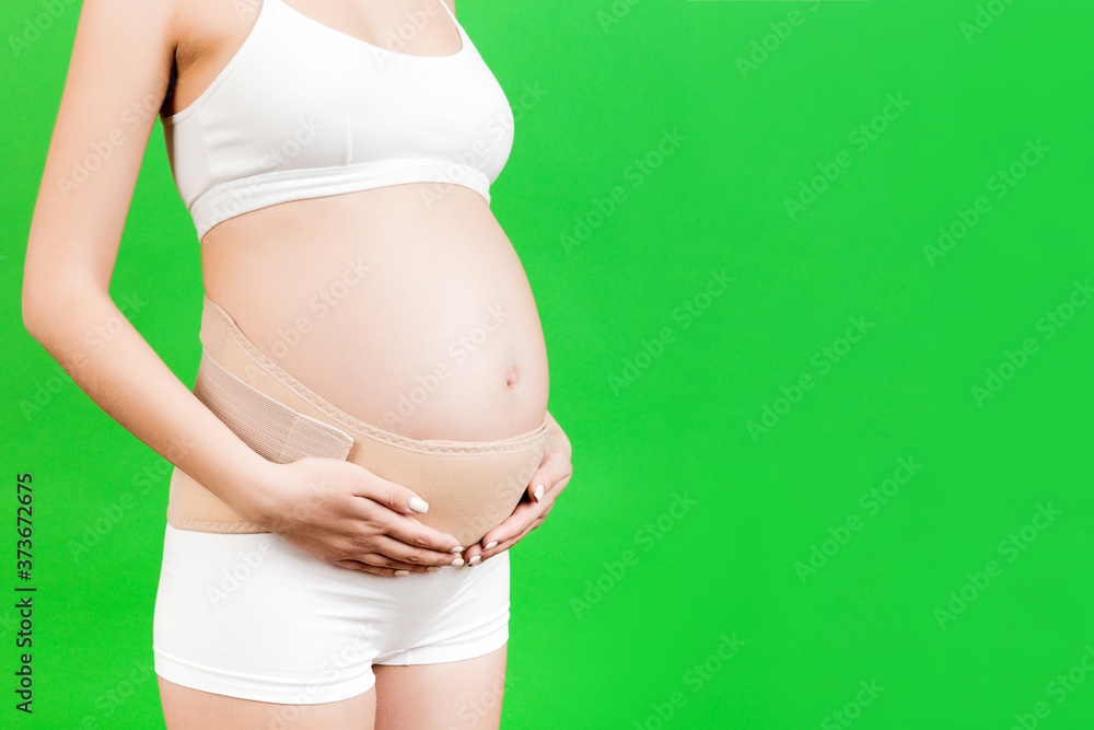 Close up of pregnant woman in underwear wearing elastic pregnancy bandage to make the pain go away at green background with copy space. Orthopedic abdominal support belt concept Close up of pregnant woman in underwear wearing elastic pregnancy bandage to make the pain go away at green background with copy space. Orthopedic abdominal support belt concept