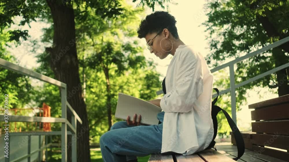 A side view a beautiful young african american woman is reading a book sitting on the bench in the park A side view a beautiful young african american woman is reading a book sitting on the bench in the park