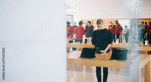 Φωτογραφία Young female customer standing near table with electronic products while examining the goods, store employees and visitors stand in blur on background