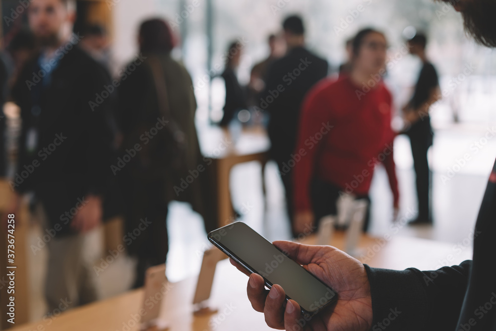 custom made wallpaper toronto digitalYoung male customer holding in hand mobile phone while examining digital goods, store employees and visitors stand in blur on background. Millennial hipster guy checking device for buy
