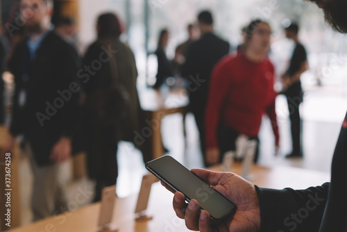 Wallpaper Mural Young male customer holding in hand mobile phone while examining digital goods, store employees and visitors stand in blur on background. Millennial hipster guy checking device for buy Torontodigital.ca