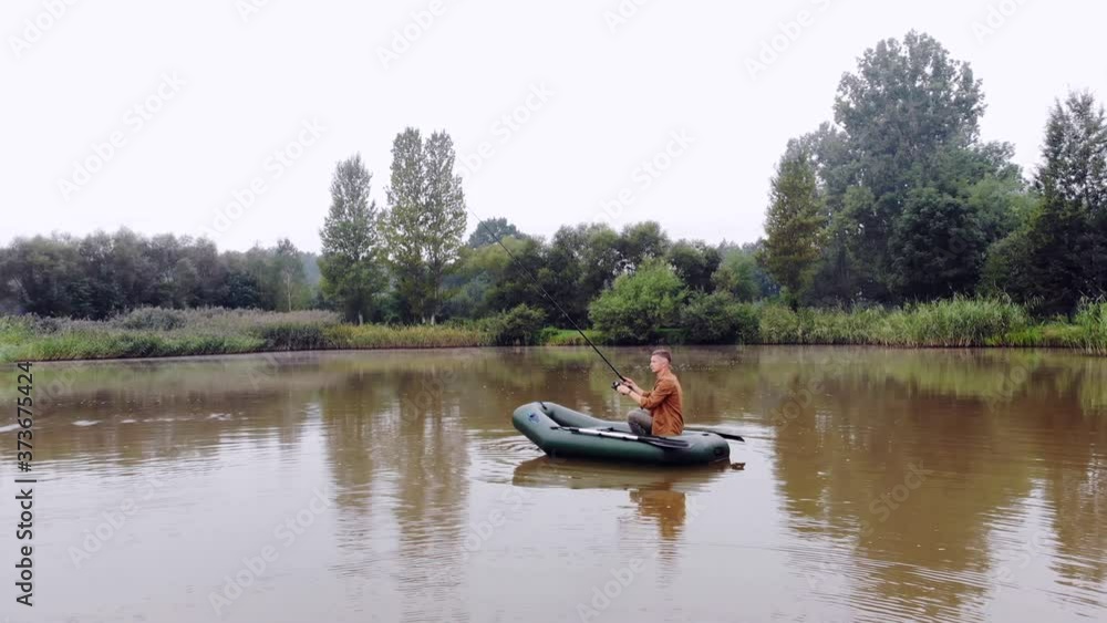 young man on an inflatable boat sits and throws a spinning or fishing rod into the water in the middle a lake or river. background of reeds and forest. Hipster guy fisherman active recreation, camping young man on an inflatable boat sits and throws a spinning or fishing rod into the water in the middle a lake or river. background of reeds and forest. Hipster guy fisherman active recreation, camping