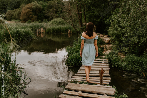 A tanned brunette girl in a turquoise dress with white polka dots walks across the river alone an old wooden bridge. Against background of dark green trees. cloudy weather. back view. Woman at nature
