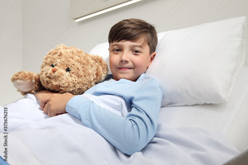 Portrait of cute happy child alone in hospital room lying in bed with teddy bear looking at camera and smiling with copy space Portrait of cute happy child alone in hospital room lying in bed with teddy bear looking at camera and smiling with copy space