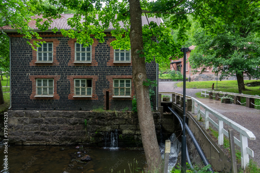 Fiskars village and its mill in the summer time. Former iron works mill built of slag brick (built in 1898). Some trees and a river, Finland. Fiskars village and its mill in the summer time. Former iron works mill built of slag brick (built in 1898). Some trees and a river, Finland.