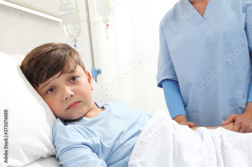 Portrait of sad child lying in bed in hospital room with nurse behind him holding his harm