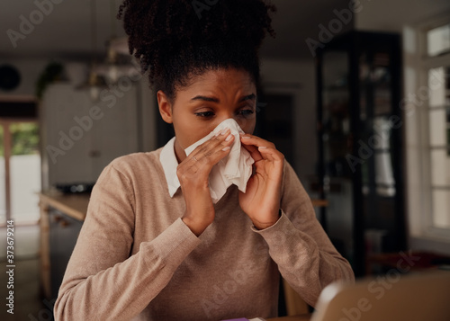 Valokuva Sick woman blowing nose in tissue paper while sitting at home using laptop