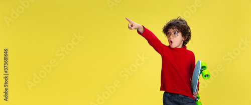 Pointing. Portrait of pretty young curly boy in red wear on yellow studio background. Childhood, expression, education, fun concept. Preschooler with bright facial expression and sincere emotions